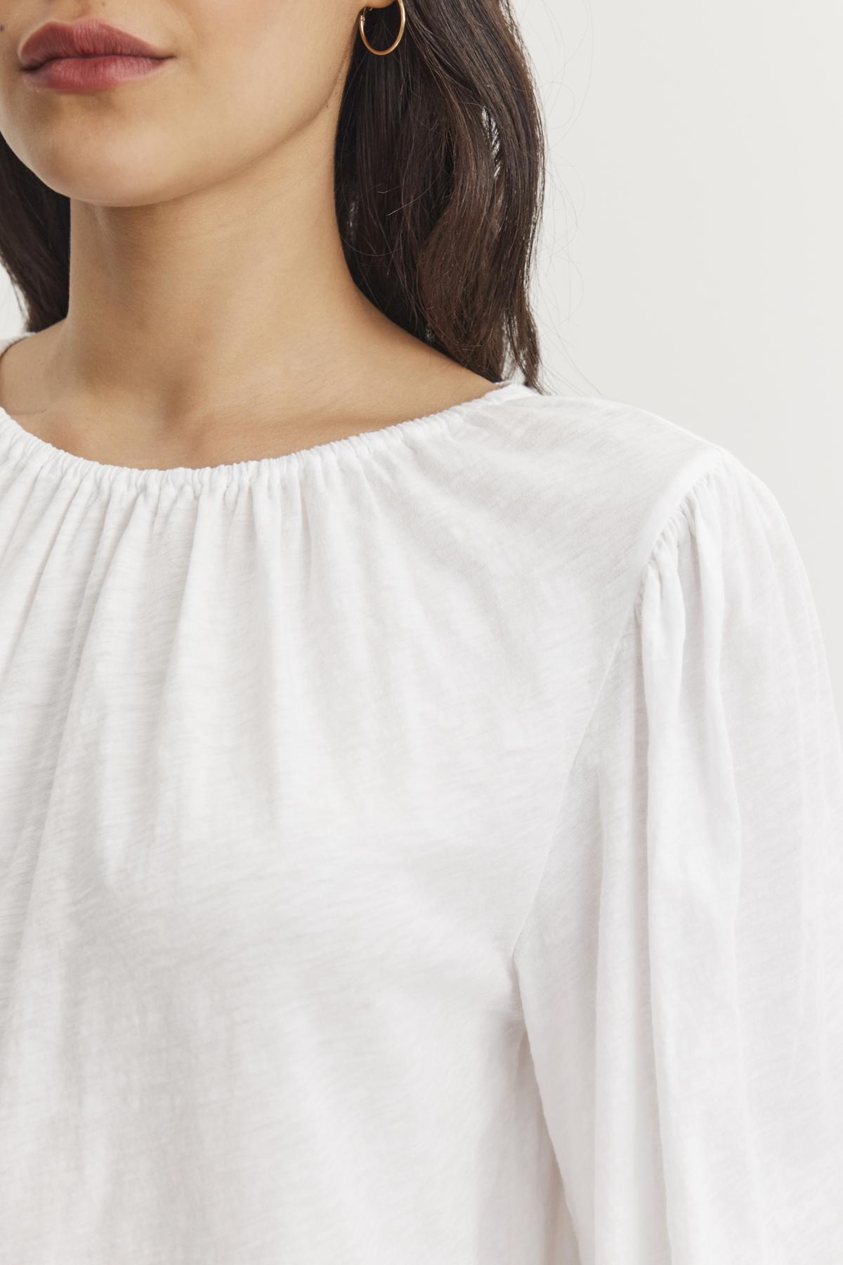A person is wearing the CARENA TEE from Velvet by Graham & Spencer, a white cotton slub blouse with an elastic neckline and puffed sleeves, visible from the shoulders up, paired with gold hoop earrings.
