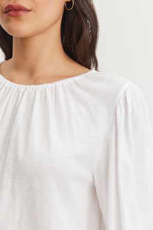 A person is wearing the CARENA TEE from Velvet by Graham & Spencer, a white cotton slub blouse with an elastic neckline and puffed sleeves, visible from the shoulders up, paired with gold hoop earrings.