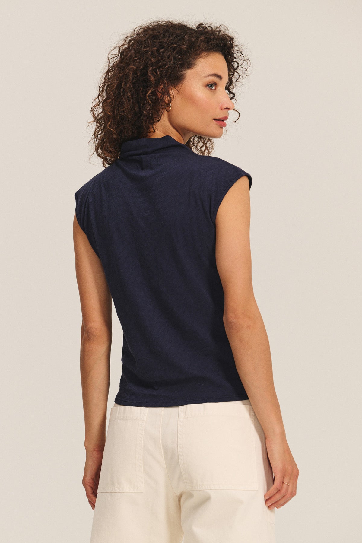 A woman with curly hair, wearing the navy FLETCHER TEE by Velvet by Graham & Spencer and white lightweight cotton slub pants, stands facing away from the camera against a plain background.-40333264617665