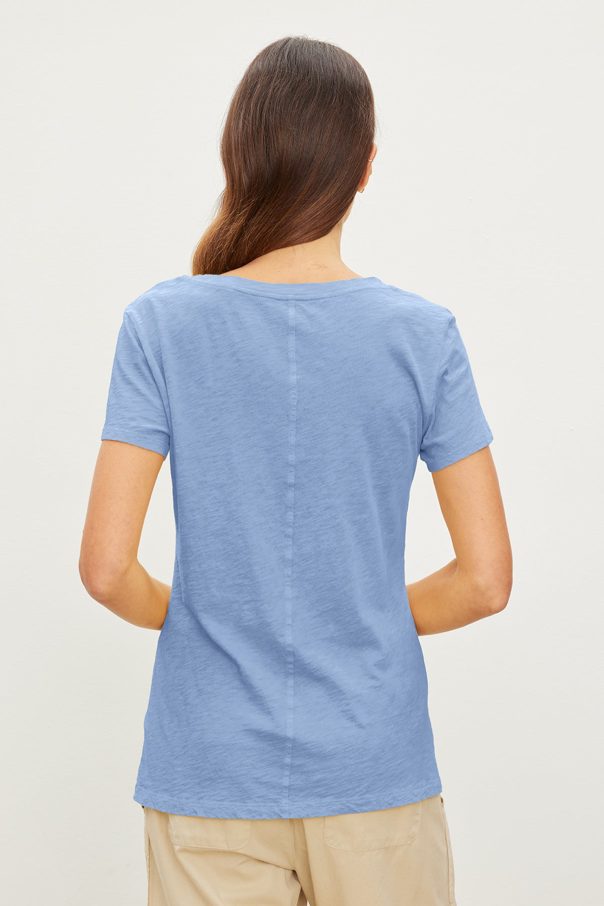 A person with long brown hair is wearing a light blue, flattering fit, short-sleeve V-neck LILITH TEE by Velvet by Graham & Spencer and beige pants, shown from the back against a plain light-colored background.-35967729008833