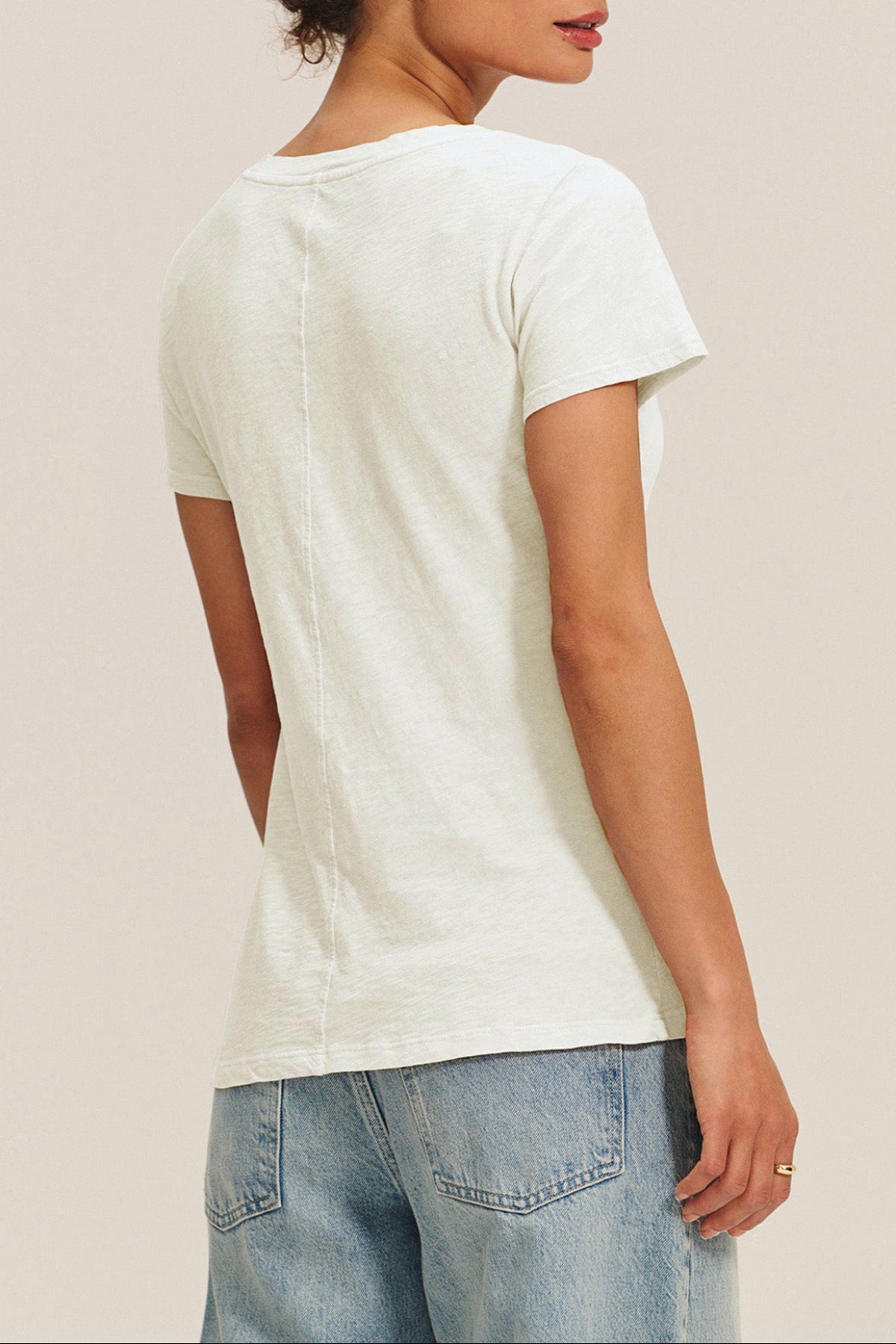 A woman shown from the back wears the Velvet by Graham & Spencer LILITH TEE, a plain white cotton slub V-neck, paired with light blue denim jeans against a neutral background.-41637039734977