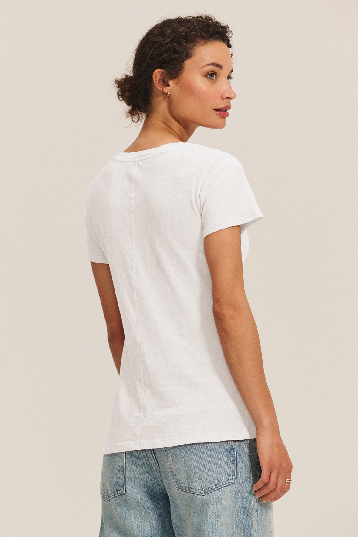 A woman stands turned to the side, looking over her shoulder, wearing the LILITH TEE by Velvet by Graham & Spencer and light blue jeans against a plain background.-41491726205121