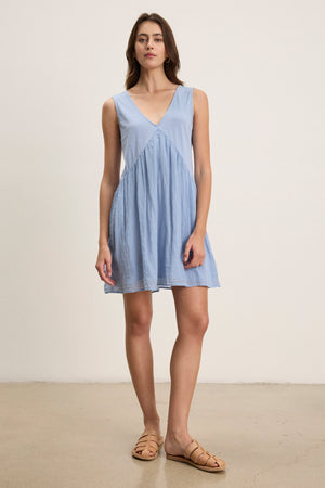 A woman stands indoors against a plain background wearing the LILY DRESS by Velvet by Graham & Spencer—a sleeveless, light blue V-neck mini dress—paired with tan sandals.