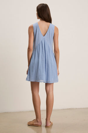 A person stands indoors with their back turned, wearing the LILY DRESS by Velvet by Graham & Spencer—a light blue mini dress featuring a deep V-back and V-neckline—paired with tan sandals against a plain background.