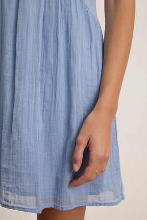 A person models the LILY DRESS by Velvet by Graham & Spencer—a light blue, textured mini dress with a V-neckline—standing with one arm relaxed, displaying a gold ring on their finger.