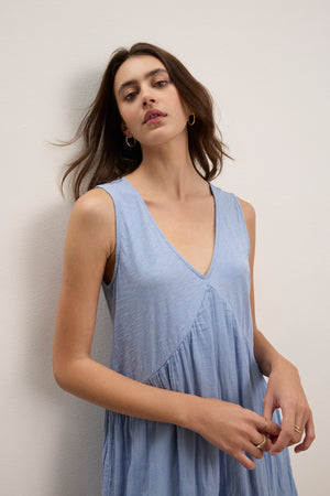 A woman with medium-length brown hair wears the LILY DRESS by Velvet by Graham & Spencer—a sleeveless, light blue V-neck mini dress—while standing against a plain light-colored wall.
