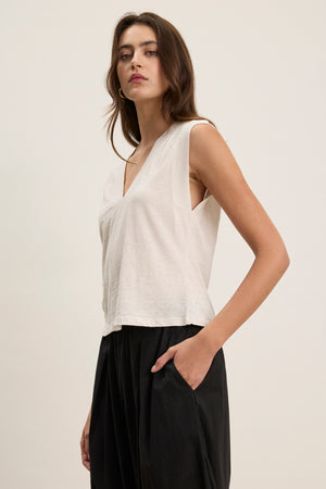 A woman stands against a plain background wearing the MARIA TEE by Velvet by Graham & Spencer, a white sleeveless V-neck top in lightweight, breathable cotton slub, paired with black pants and one hand in her pocket.
