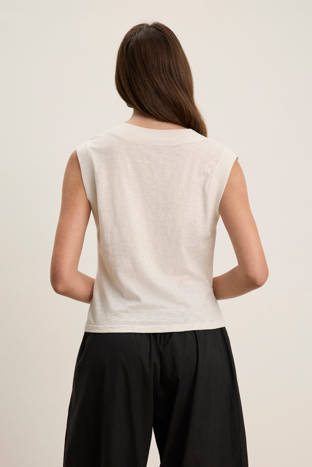 A person with long brown hair, wearing the MARIA TEE by Velvet by Graham & Spencer—a white sleeveless boxy cropped top—and black pants, stands with their back to the camera against a plain background.-39120069198017