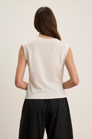 A person with long brown hair, wearing the MARIA TEE by Velvet by Graham & Spencer—a white sleeveless boxy cropped top—and black pants, stands with their back to the camera against a plain background.