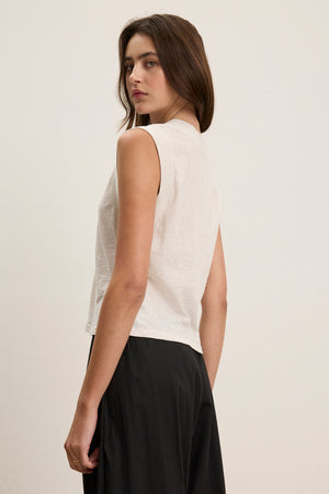 A woman with long brown hair wears the MARIA TEE by Velvet by Graham & Spencer—a sleeveless white top in lightweight breathable textured cotton slub—with a black skirt, standing sideways against a plain light background.
