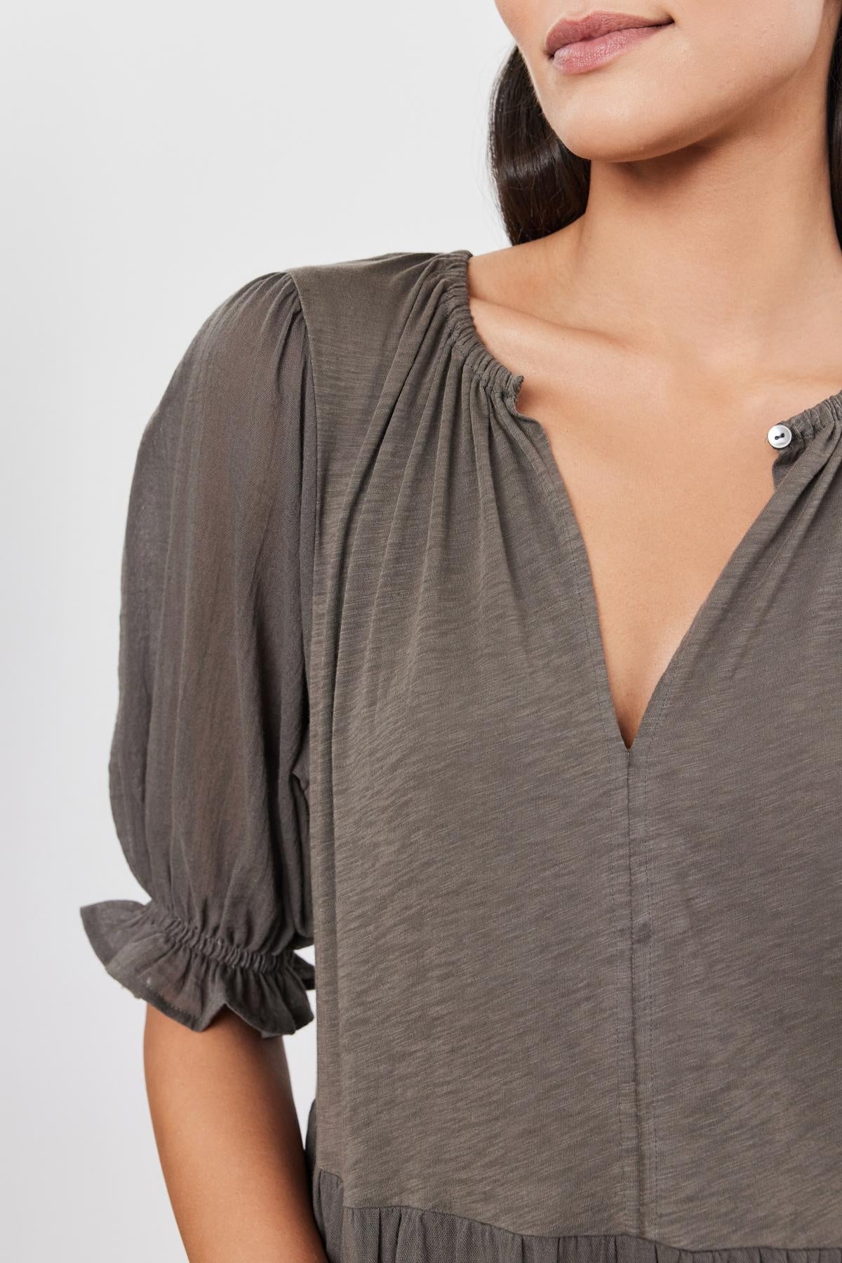 A woman is wearing the LEIGHTON DRESS by Velvet by Graham & Spencer. The dress features a grey, long-sleeve top with a gathered neckline and a deep V-cut, accented by airy cotton gauze sleeves that add an elegant touch. The focus is on the upper part of her torso.-37575436697793