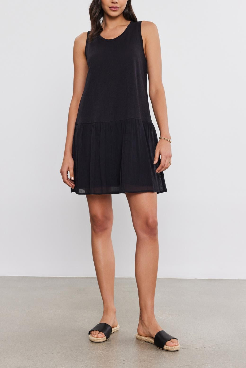 A woman in a sleeveless black Velvet by Graham & Spencer MINA DRESS with a tiered skirt and black sandals stands on a plain white background.-37054552375489