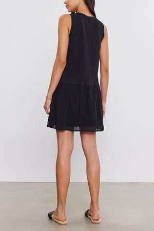 A woman wearing a sleeveless black MINA DRESS by Velvet by Graham & Spencer with a tiered skirt stands with her back to the camera, in a simple, well-lit studio setting.