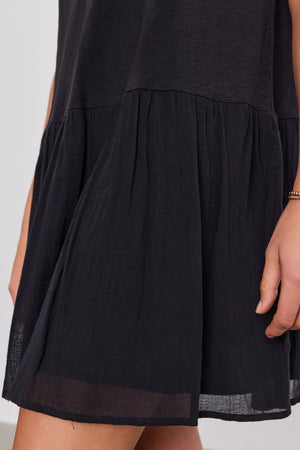 Close-up of a section of a black sleeveless MINA DRESS with a tiered, gathered skirt made from cotton slub by Velvet by Graham & Spencer, and a woman's arm visible to the right.