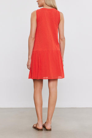 Woman standing facing away from the camera, wearing a red sleeveless Velvet by Graham & Spencer MINA DRESS with a tiered skirt and brown flat shoes, on a plain background.