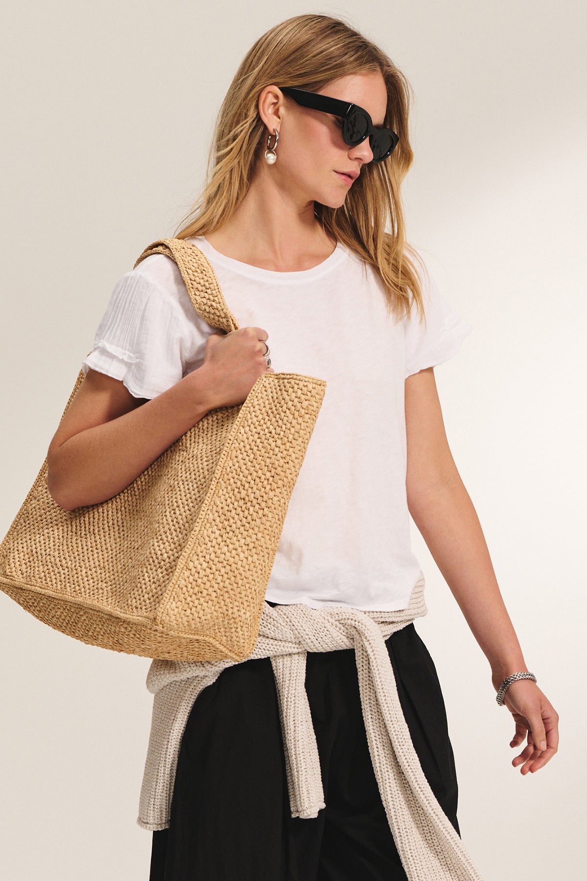 Woman wearing black sunglasses, a white t-shirt, black pants, and the Velvet by Graham & Spencer THALIA CARDIGAN with long sleeves tied at the waist, holding a large woven straw tote bag.-39292671852737