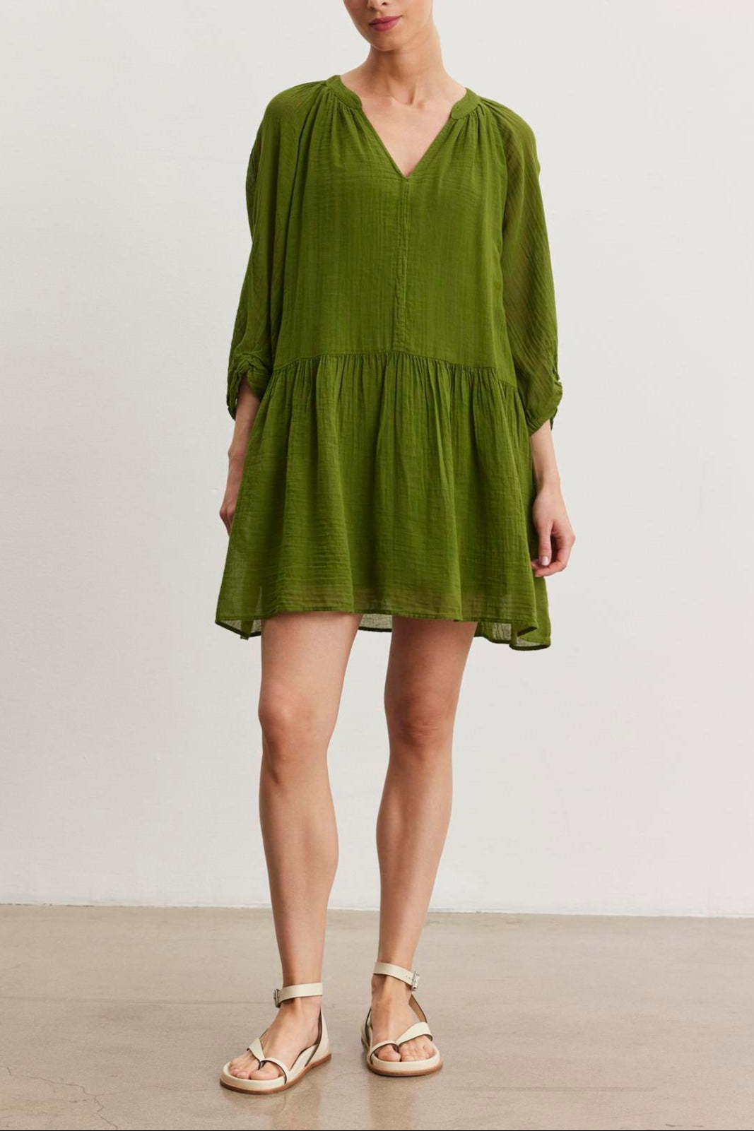 A person stands facing forward, wearing a loose-fitting green V-neck Sloan Dress from Velvet by Graham & Spencer, made from breathable cotton gauze, paired with beige sandals. The background is a plain, light-colored wall.-37575450558657