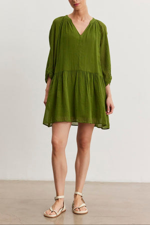 A person stands facing forward, wearing a loose-fitting green V-neck Sloan Dress from Velvet by Graham & Spencer, made from breathable cotton gauze, paired with beige sandals. The background is a plain, light-colored wall.