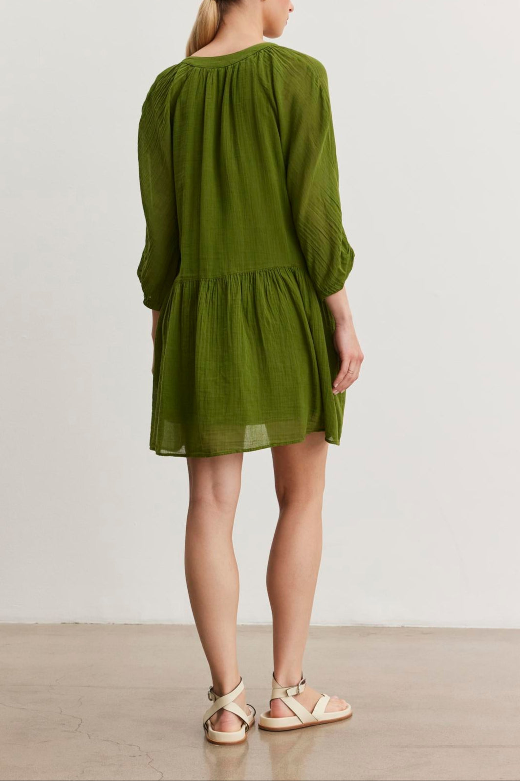 A woman with blonde hair is standing with her back to the camera, wearing the versatile SLOAN DRESS from Velvet by Graham & Spencer, a green long-sleeved dress made of breathable cotton gauze. She is also wearing white sandals.-37575450591425