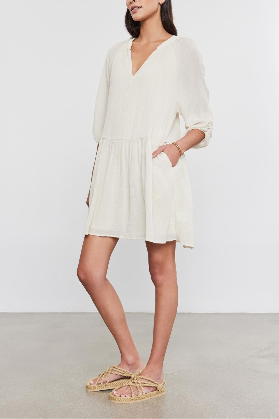 A woman in a white Velvet by Graham & Spencer Sloan Dress with pleat detailing and beige sandals stands against a gray background, looking to the side with her hands slightly behind her.