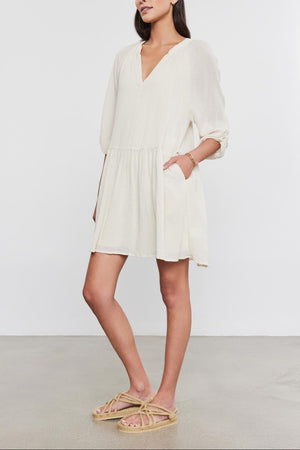 A woman in a white Velvet by Graham & Spencer Sloan Dress with pleat detailing and beige sandals stands against a gray background, looking to the side with her hands slightly behind her.