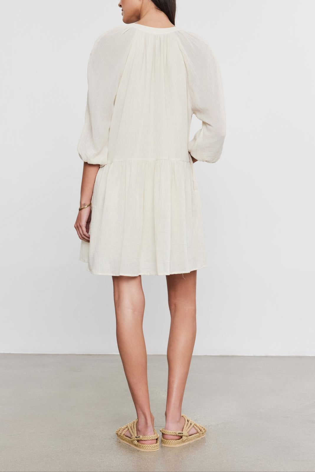 A woman standing and facing away from the camera, wearing a short, pale beige Velvet by Graham & Spencer Sloan Dress with pleat detailing, puffed sleeves, and open-toe sandals.