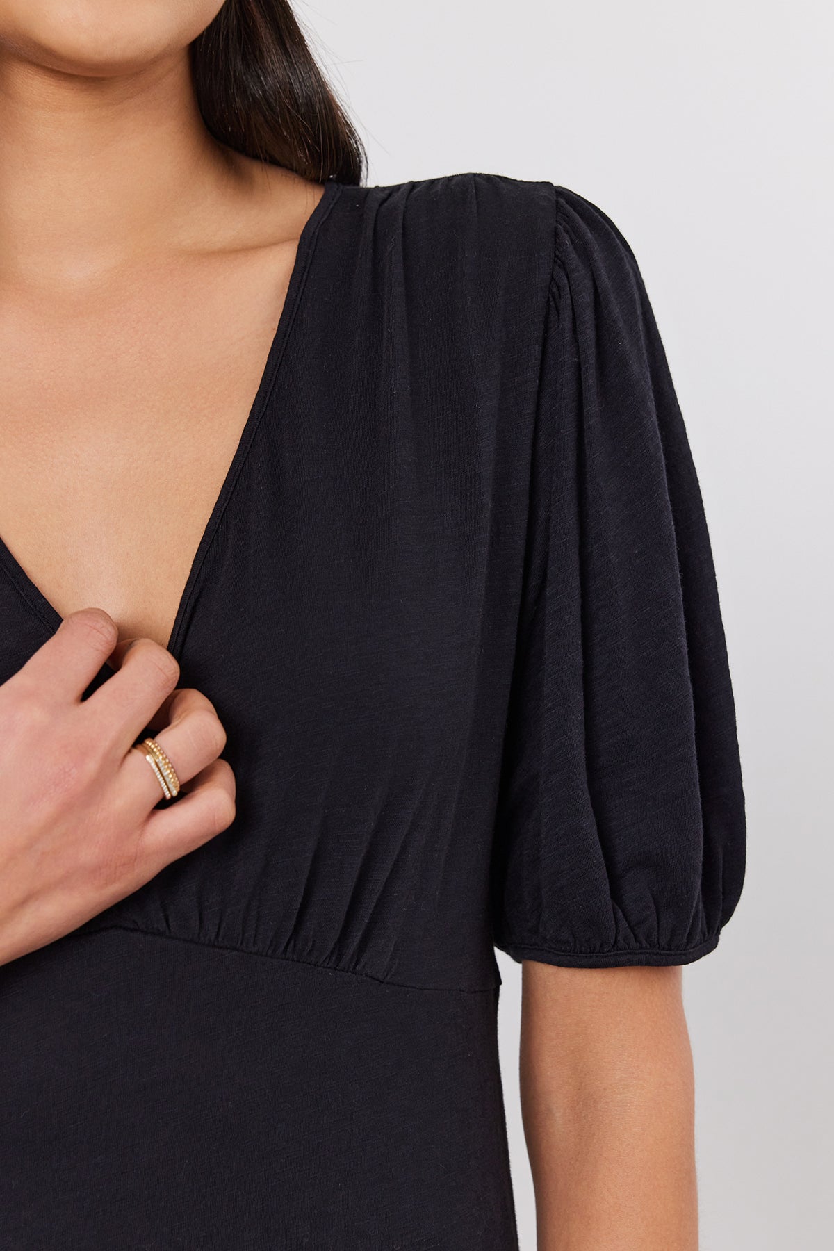 A person wearing a dark MORGAN DRESS by Velvet by Graham & Spencer, possibly made of cotton slub, holds the fabric at their chest with one hand, adorned with a gold ring. The background is plain white.-37452675678401