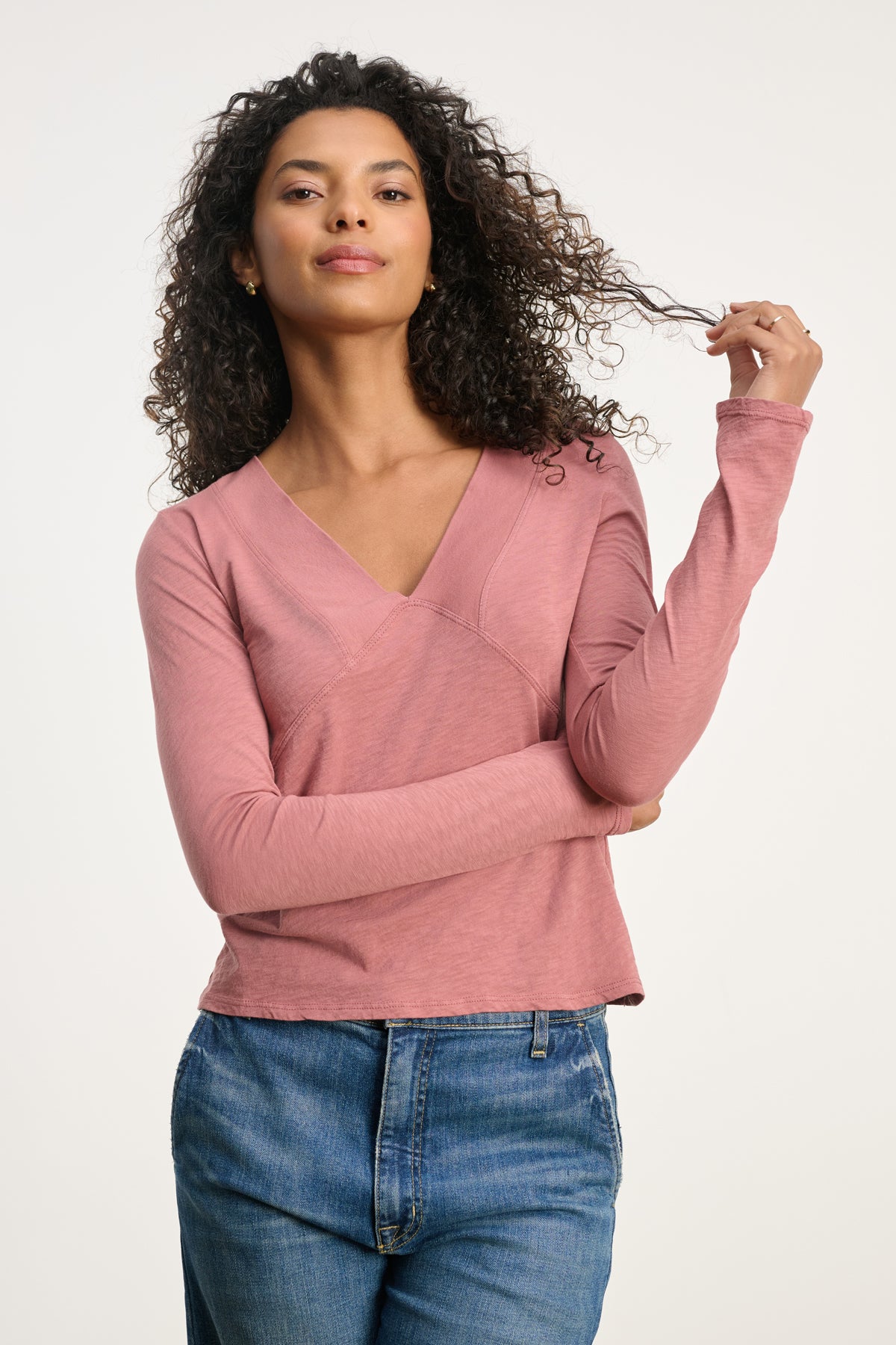 A woman with curly hair, dressed in the Velvet by Graham & Spencer NESSA TEE—a long-sleeve deep V-neck—paired with blue jeans, stands against a plain background, holding a strand of her hair.-39838366924993