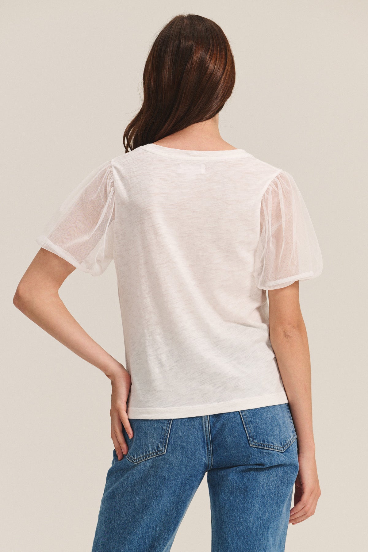 A person stands facing away, wearing the JUSTINE TEE by Velvet by Graham & Spencer—a white cotton slub top with tulle puff sleeves—paired with blue jeans against a plain background.-40916286865601