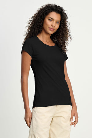 A woman with curly hair wears the ODELIA TEE by Velvet by Graham & Spencer, a premium cotton crewneck, paired with light-colored pants against a neutral background.