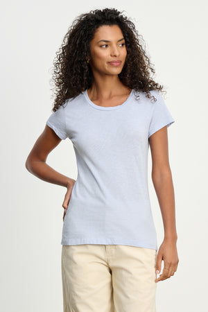 A woman with curly hair wears the ODELIA TEE by Velvet by Graham & Spencer, a light blue premium cotton crewneck, paired with beige pants against a plain light background.