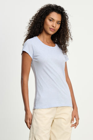A woman with curly hair wears the Velvet by Graham & Spencer ODELIA TEE, a premium cotton light blue crewneck, paired with beige pants against a plain light background.