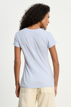 A woman with curly hair models the Velvet by Graham & Spencer ODELIA TEE in light blue, featuring a classic crew neckline, paired with cream pants and photographed from the back against a plain background.