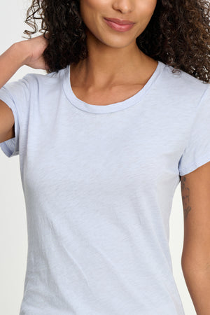 A woman with curly hair wears the ODELIA TEE by Velvet by Graham & Spencer—a light blue premium cotton top—standing with one hand behind her neck, partially revealing a tattoo on her upper arm.