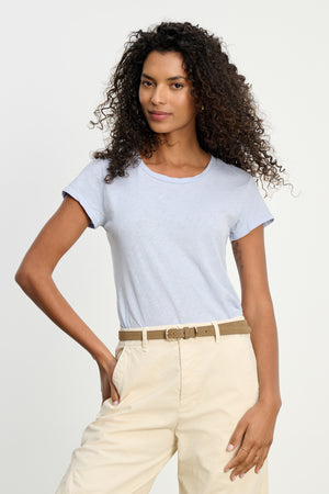 A woman with curly hair wears the ODELIA TEE by Velvet by Graham & Spencer—a light blue premium cotton crewneck—tucked into beige pants with a tan belt, standing against a plain white background.