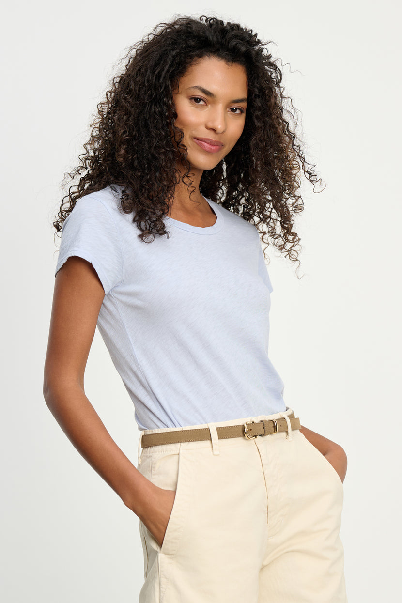 A woman with curly hair wears the ODELIA TEE by Velvet by Graham & Spencer—premium cotton, classic crew neck, light blue—paired with beige pants, hands in pockets, standing against a plain light background.