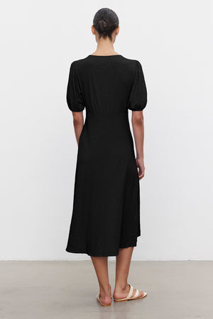 Wearing the PARKER DRESS from Velvet by Graham & Spencer, a person stands on a plain floor in tan sandals. The black, midi-length dress with short sleeves enhances their form as they face away.