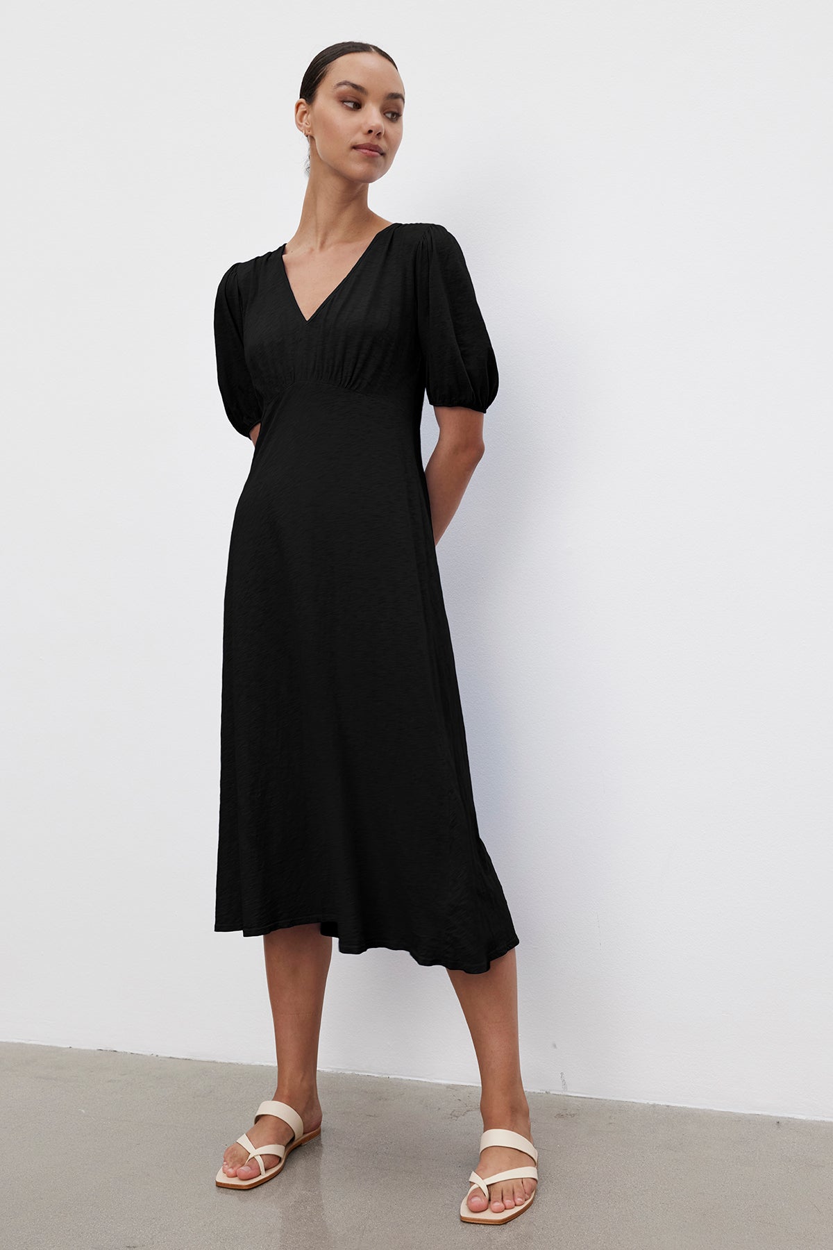 A person wearing the PARKER DRESS by Velvet by Graham & Spencer stands against a white background. The black cotton slub V-neck dress features puffed sleeves and a midi length, while their white sandals complete the look. The figure-skimming fit elegantly enhances their silhouette.-37185421902017
