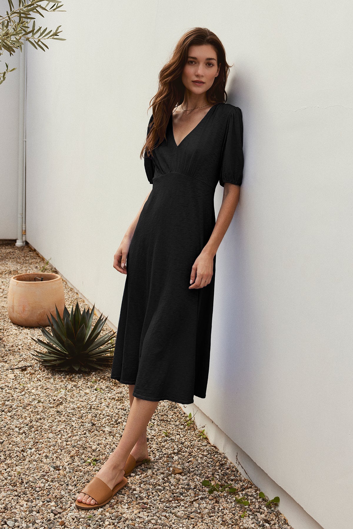 A woman in a black PARKER DRESS by Velvet by Graham & Spencer leans against a white wall in a gravel yard adorned with plants and a clay pot.-37185421967553