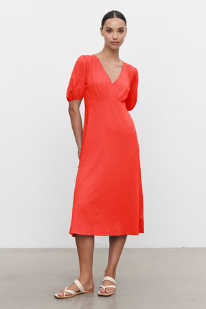 Against a plain white background, someone wears Velvet by Graham & Spencer's PARKER DRESS in bright orange with cotton slub fabric; it features a figure-skimming midi cut, short sleeves, and a V-neck, paired with flat sandals.
