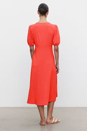 A person wears the orange PARKER DRESS by Velvet by Graham & Spencer, midi-length with a figure-skimming fit, paired with sandals, viewed from behind against a plain white background.
