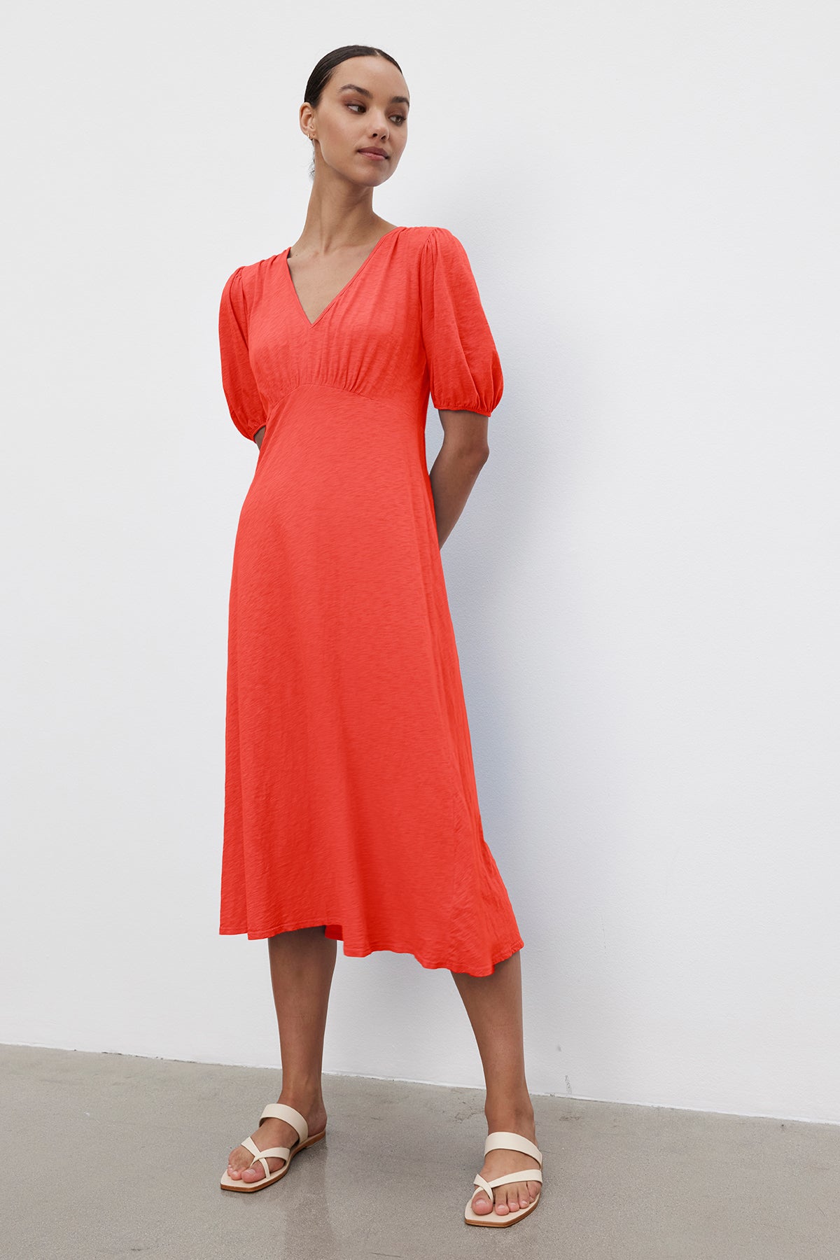 A person stands against a plain white wall, wearing a Velvet by Graham & Spencer PARKER COTTON SLUB MIDI DRESS with puffed sleeves and white sandals.-37185421869249
