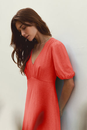 A person with long hair wearing the PARKER DRESS by Velvet by Graham & Spencer, a midi-length red cotton slub creation that offers a figure-skimming fit, stands against a white wall, gazing down slightly.