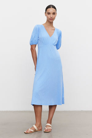 A person models Velvet by Graham & Spencer's PARKER DRESS, a light blue cotton slub with puff sleeves, figure-skimming fit, and knee-length cut. They pair it elegantly with beige sandals against a white background.