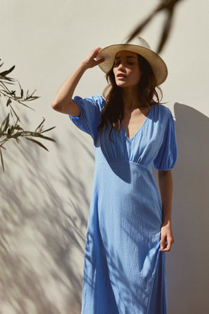 Wearing a PARKER DRESS by Velvet by Graham & Spencer, a person in blue poses against a light wall with leaf shadows, complemented by a straw hat.