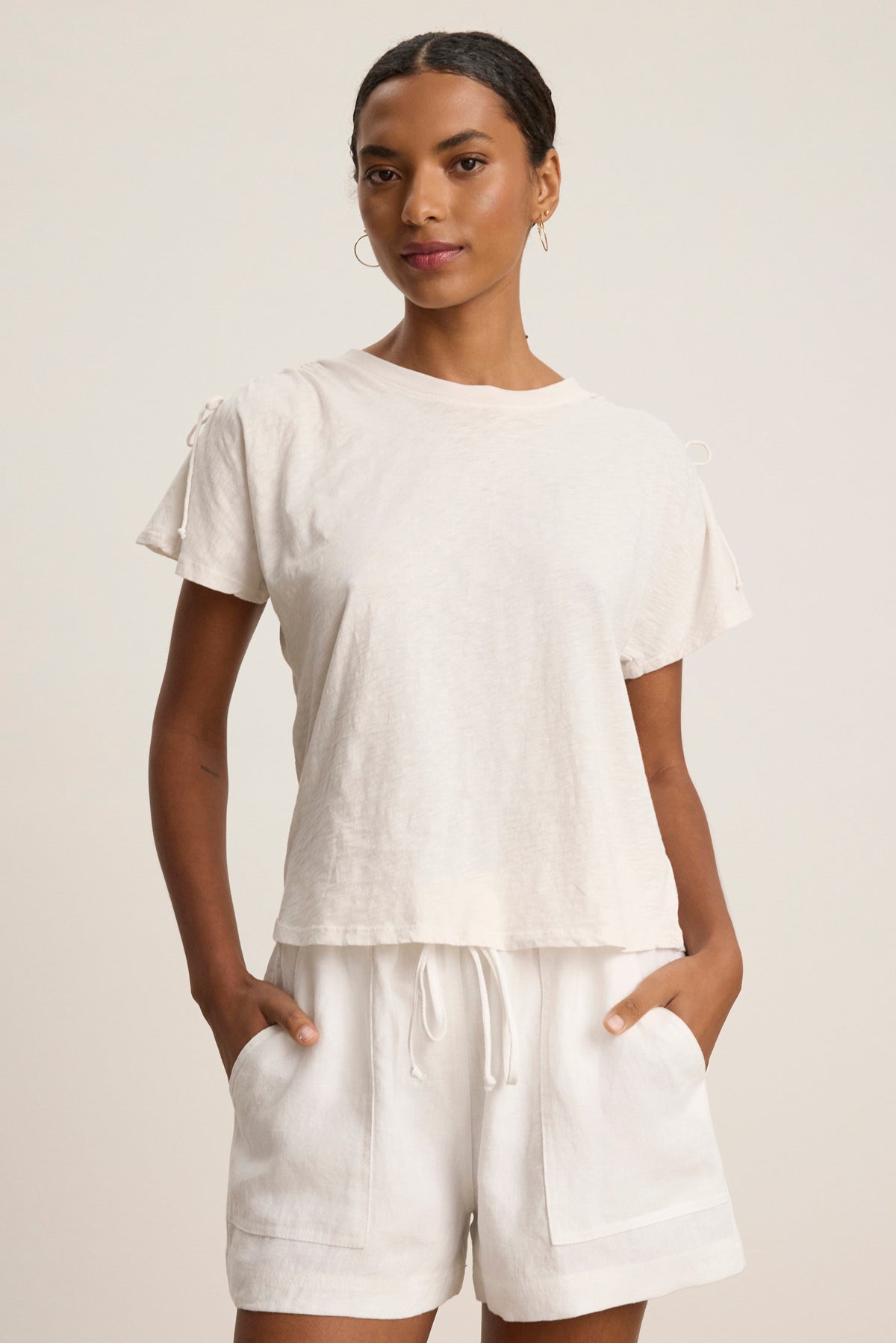 A woman stands facing forward wearing the PAULA TEE by Velvet by Graham & Spencer, a light-colored cotton slub crew-neck, paired with matching drawstring shorts, her hands in her pockets.