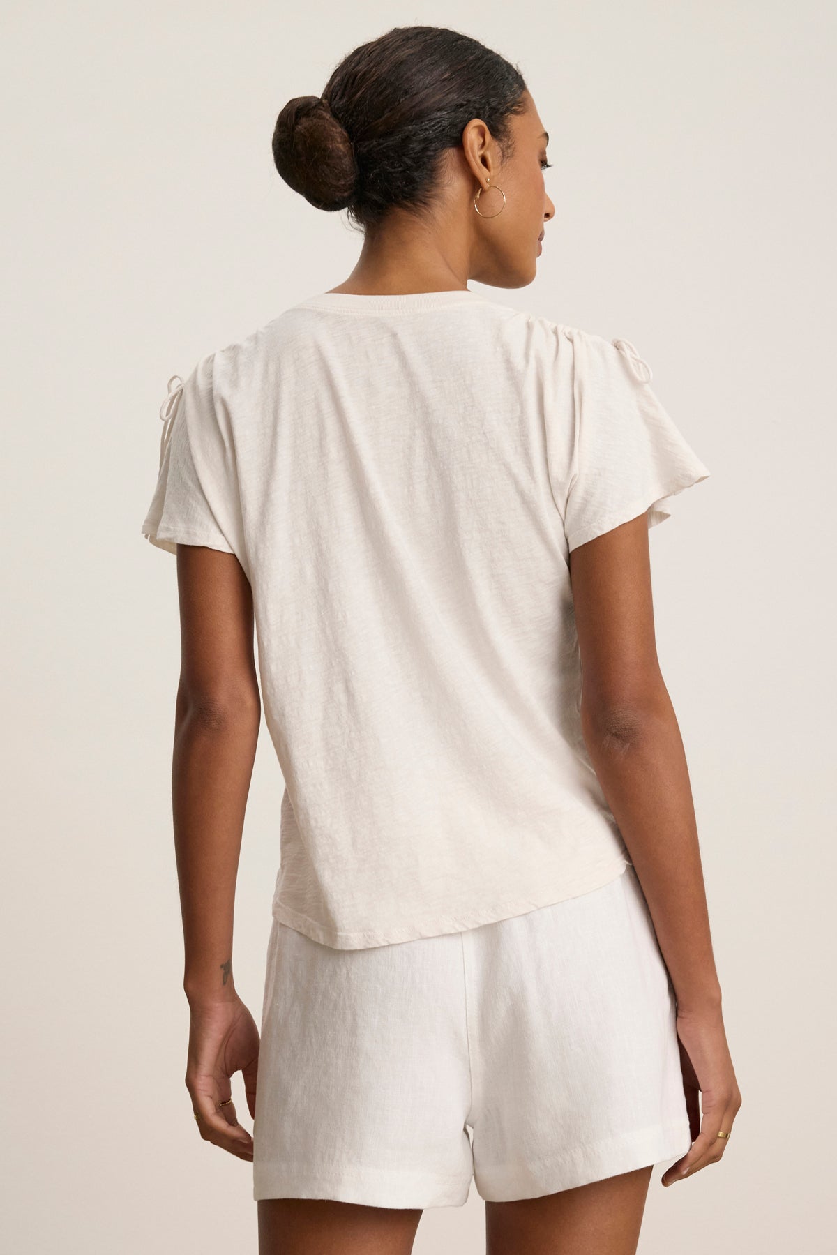 A woman faces away, hair in a bun, wearing the PAULA TEE by Velvet by Graham & Spencer— a light cotton slub crew-neck tee—paired with matching shorts against a neutral background.