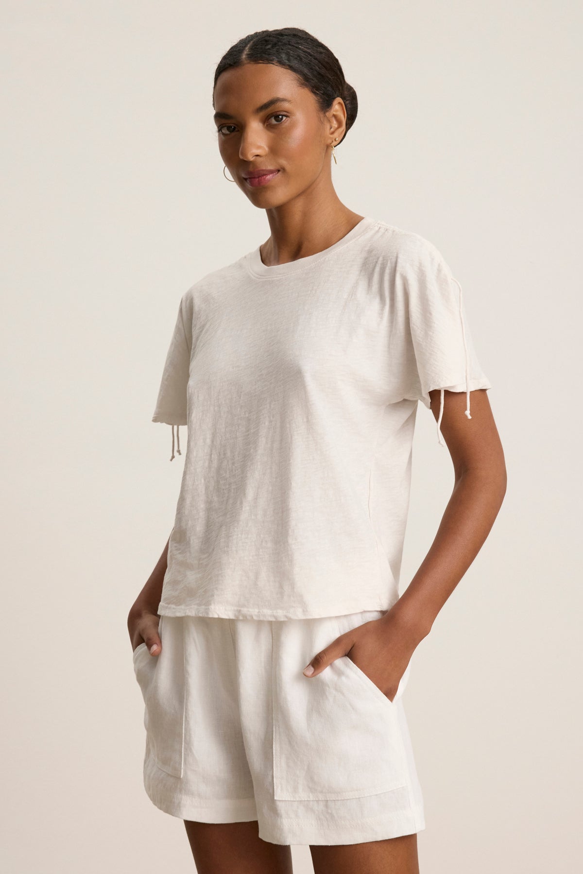 A person stands with hands in pockets, wearing the light-colored PAULA TEE by Velvet by Graham & Spencer and matching shorts against a plain background.
