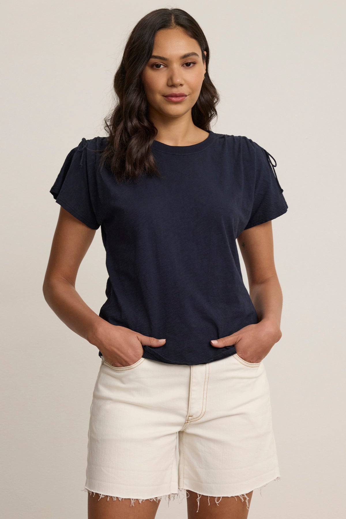 A woman stands against a plain background wearing the PAULA TEE by Velvet by Graham & Spencer, a navy cotton slub crew-neck, paired with light-colored frayed hem shorts, hands in her pockets.