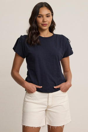 A woman stands against a plain background wearing the PAULA TEE by Velvet by Graham & Spencer, a navy cotton slub crew-neck, paired with light-colored frayed hem shorts, hands in her pockets.